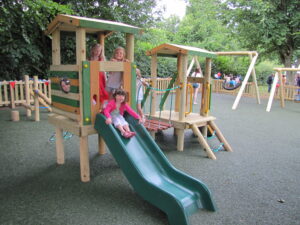 Four children play on a wooden playground structure with a slide and swings, surrounded by trees and a wooden fence, enjoying the lively atmosphere often found in public parks.