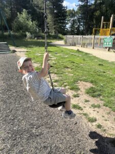 A young boy wearing a striped shirt and cap is sitting on a swing at a sunny public park playground, smiling at the camera.