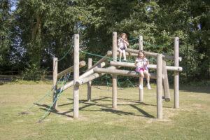 Two children sit on a wooden playground climbing frame with green ropes in a grassy outdoor area surrounded by trees, enjoying the vibrant atmosphere of public parks.
