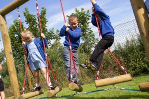 Three young children in school uniforms play on a log swing climbing frame outdoors, holding onto red ropes for balance, with grass and a fence in the background—a reminder of the importance of maintaining playground equipment for safety.