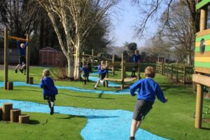 Children in blue uniforms play on a school playground with climbing apparatus, artificial grass, and trees under a clear sky, showcasing the importance of maintaining playground apparatus for safety and fun.