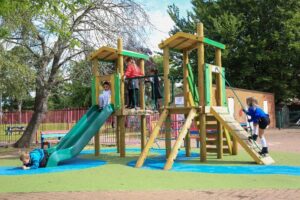 Several children play on a wooden playground structure with slides and ladders in an outdoor park area, where maintaining playground equipment ensures a safe and enjoyable space surrounded by trees and a fence.