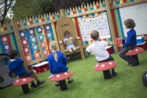Five children in a colourful outdoor classroom embody the spirit of making playgrounds more creative; one child sits on a wooden chair reading while four others sit on whimsical, mushroom-shaped stools facing them.