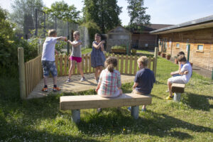 Six children are outside on a sunny day; two stand and gesture on a wooden stage while four sit on benches watching. Their imaginative scene is a testament to the importance of making playgrounds more creative, with buildings, trees, and a fence providing an inspiring backdrop.
