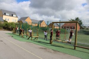 Children enjoy fun outdoor activities as they play on wooden climbing apparatus in a fenced school playground, with residential houses and cloudy skies in the background.