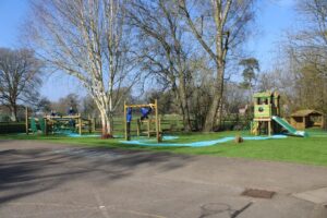Outdoor playground with wooden climbing structures, slides, and artificial grass, surrounded by bare trees. Blue tarpaulins are spread across the ground on a sunny day, offering the perfect setting for fun outdoor activities for children of all ages.