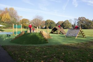 Children in red uniforms enjoy fun outdoor activities as they play on a green playground with climbing frames and grassy areas, surrounded by trees under a blue sky.