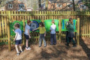 Five children interact with colourful activity boards mounted on a wooden fence in an outdoor area, engaging in fun outdoor activities amid fallen leaves and houses in the background.