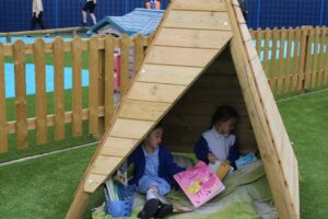 Two young girls sit inside a small wooden tent on artificial grass, enjoying Fun Outdoor Activities as they read books together. A wooden fence and blue play area are visible in the background.
