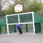 A young boy in a blue jacket stands near a green outdoor sports structure with a basketball hoop and fencing on a paved playground.