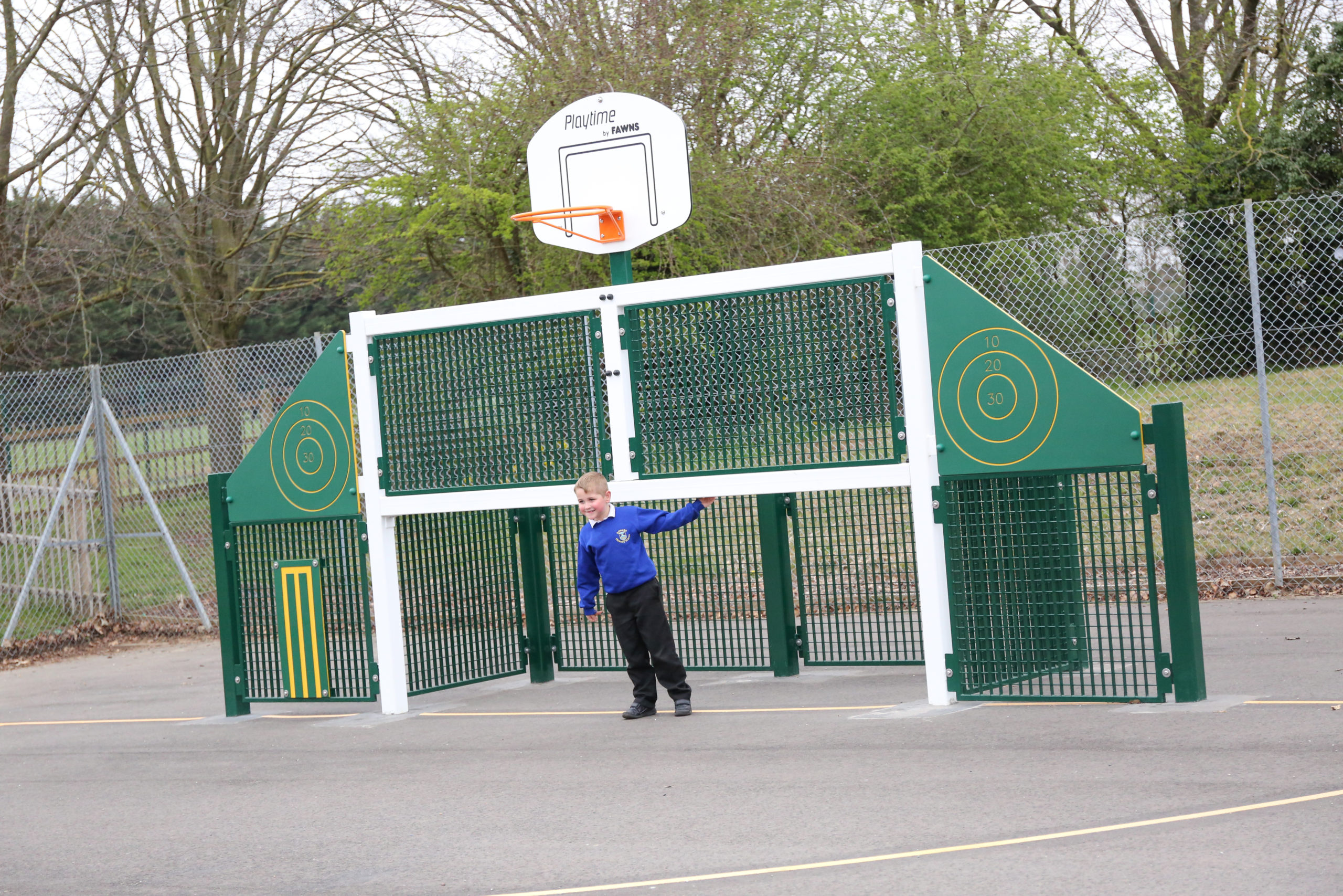 A young boy in a blue jacket stands near a green outdoor sports structure with a basketball hoop and fencing on a paved playground.