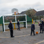 Six children in school uniforms are playing with an orange basketball on an outdoor court near a basketball net, with houses and trees in the background.