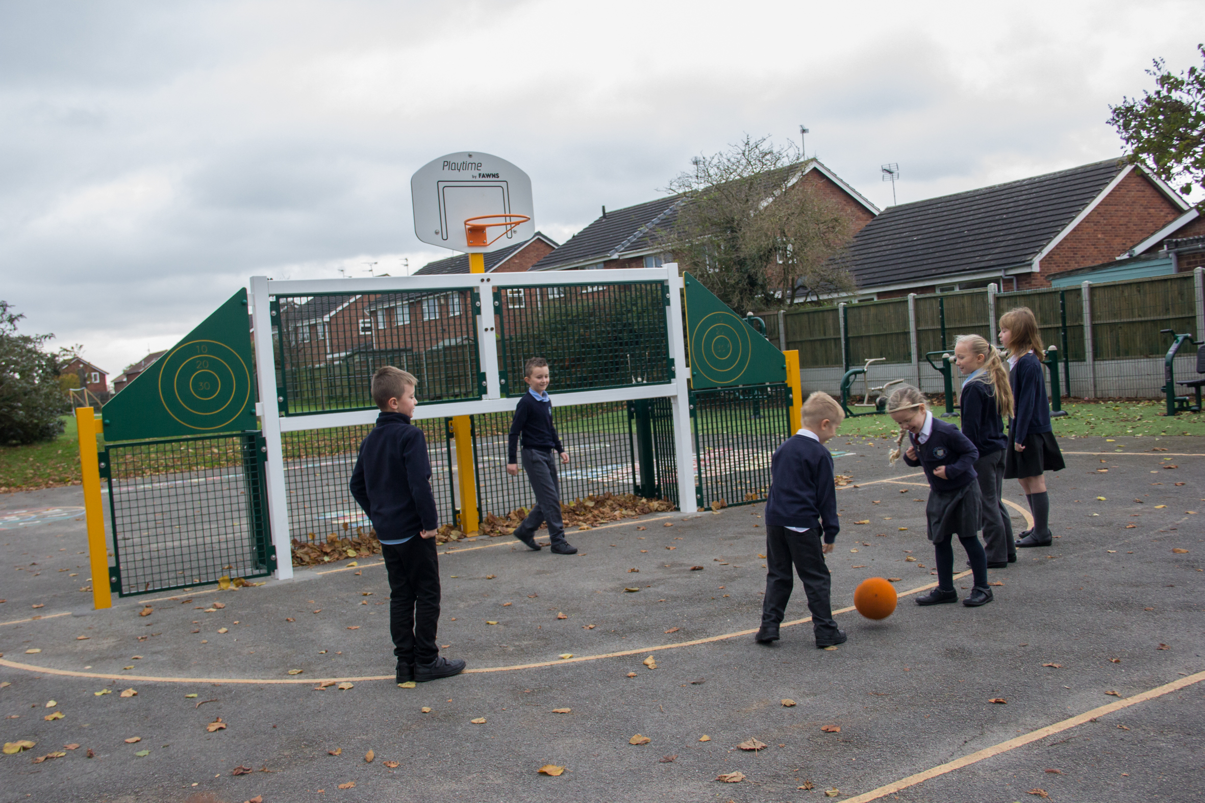 Six children in school uniforms are playing with an orange basketball on an outdoor court near a basketball net, with houses and trees in the background.