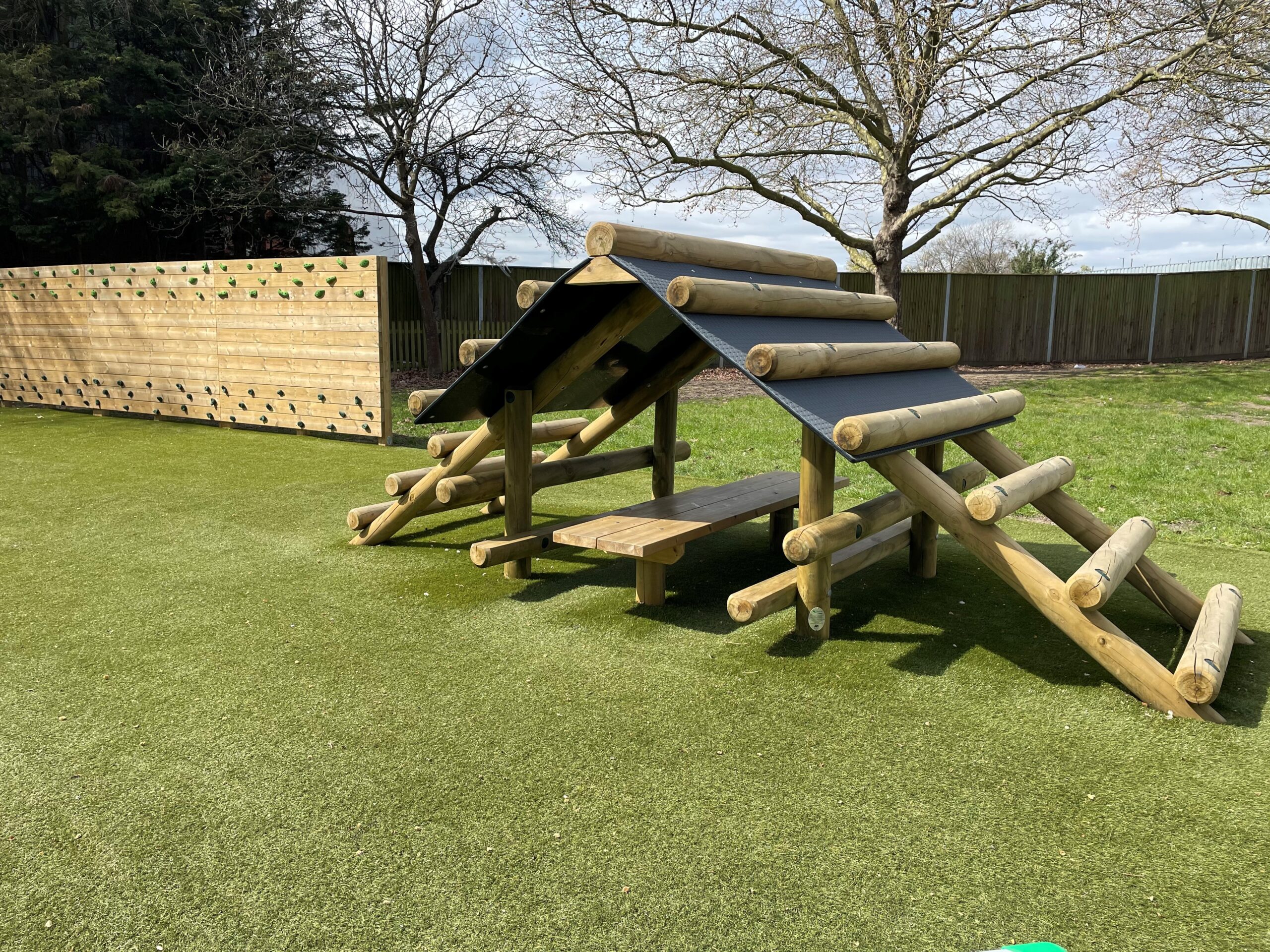 A wooden Cabin Climber with a roof and benches sits on artificial grass near a wooden climbing wall and leafless trees.