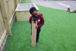 A young child in a maroon hoodie explores a Bug Magnifier Post, peering through the wooden and metal outdoor viewing device on artificial grass near a playground.