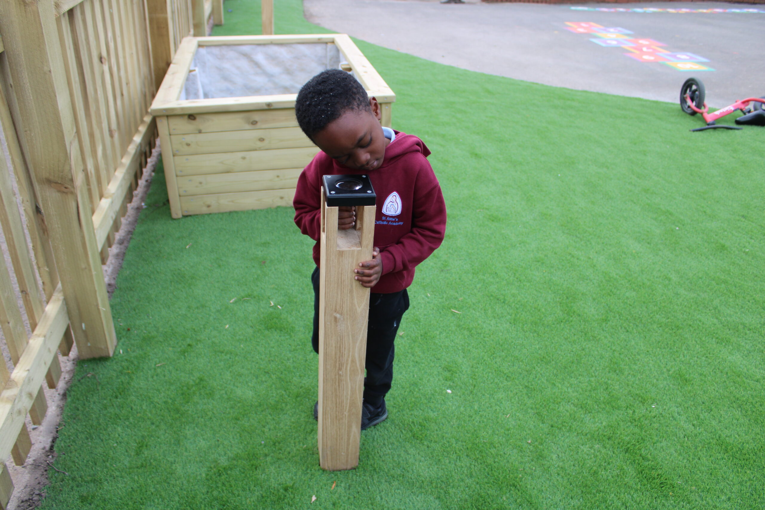 A young child in a maroon hoodie explores a Bug Magnifier Post, peering through the wooden and metal outdoor viewing device on artificial grass near a playground.