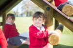 Three young children in red school uniforms play on the wooden Cabin Climber outdoors on a sunny day.