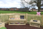 A wooden play kitchen set stands in a fenced outdoor area, featuring toy food items scattered on a messy table, with shelves and benches; houses and grass are visible in the background.