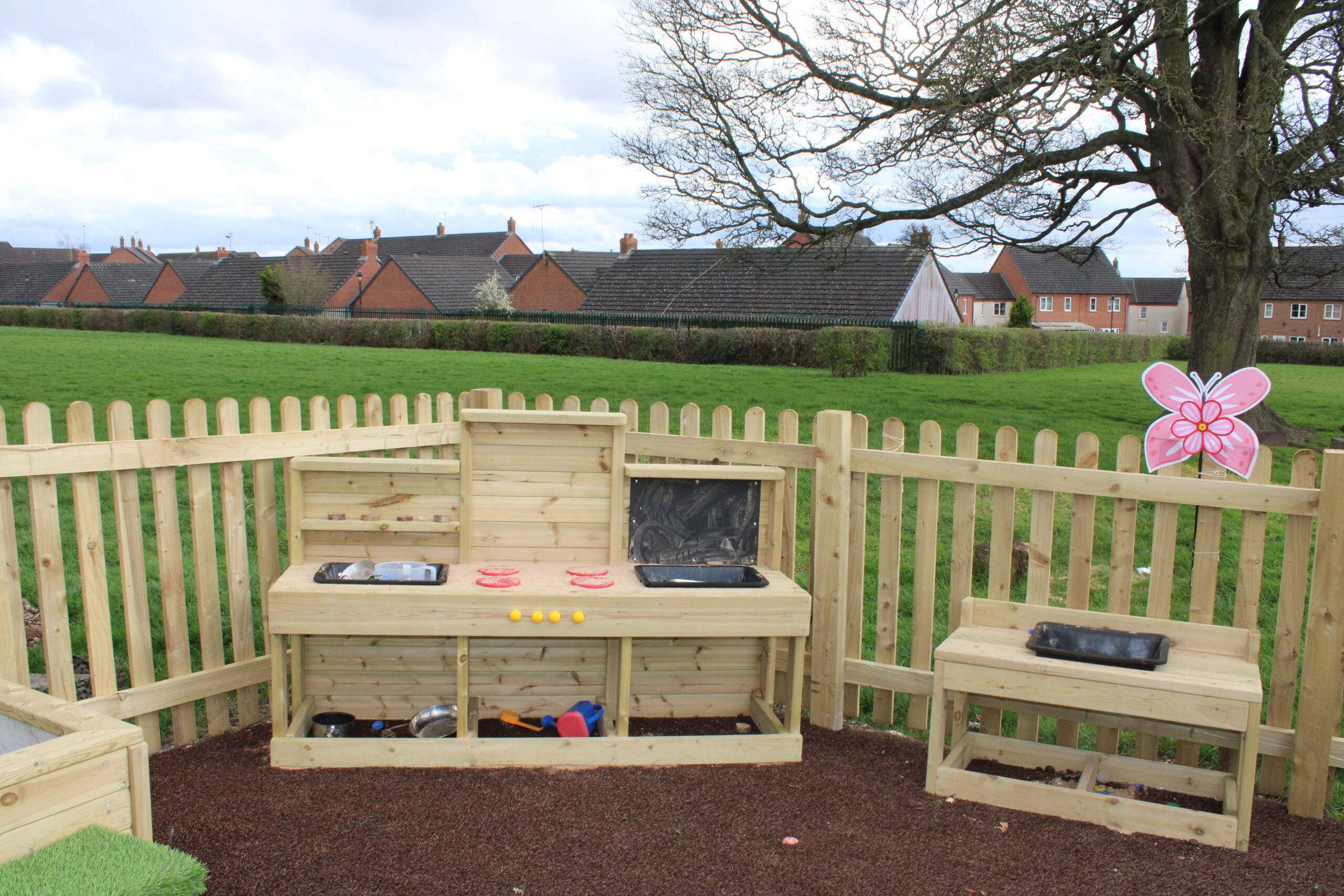 A wooden play kitchen set stands in a fenced outdoor area, featuring toy food items scattered on a messy table, with shelves and benches; houses and grass are visible in the background.