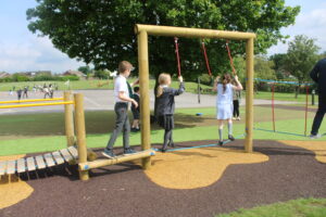 Children walk across a wooden Link Mini Balance Beam obstacle course on a school field, with other pupils playing in the background.