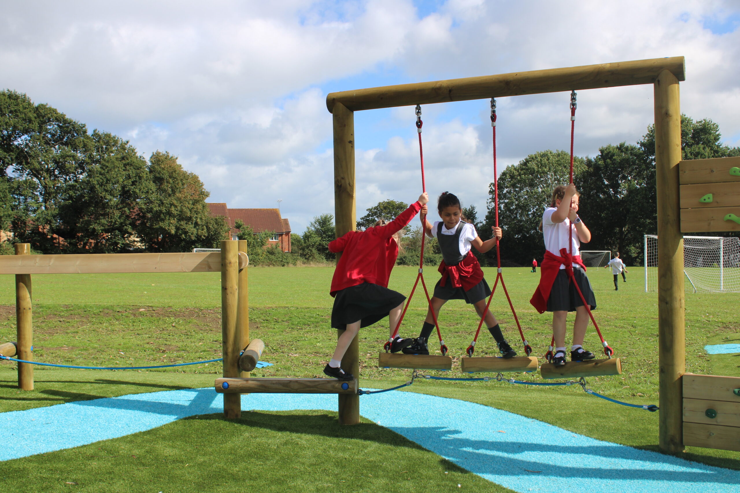 Three children in school uniforms play on a wooden rope bridge and Link Mini Balance Beam in an outdoor playground, with a grassy field and football goals visible in the background.