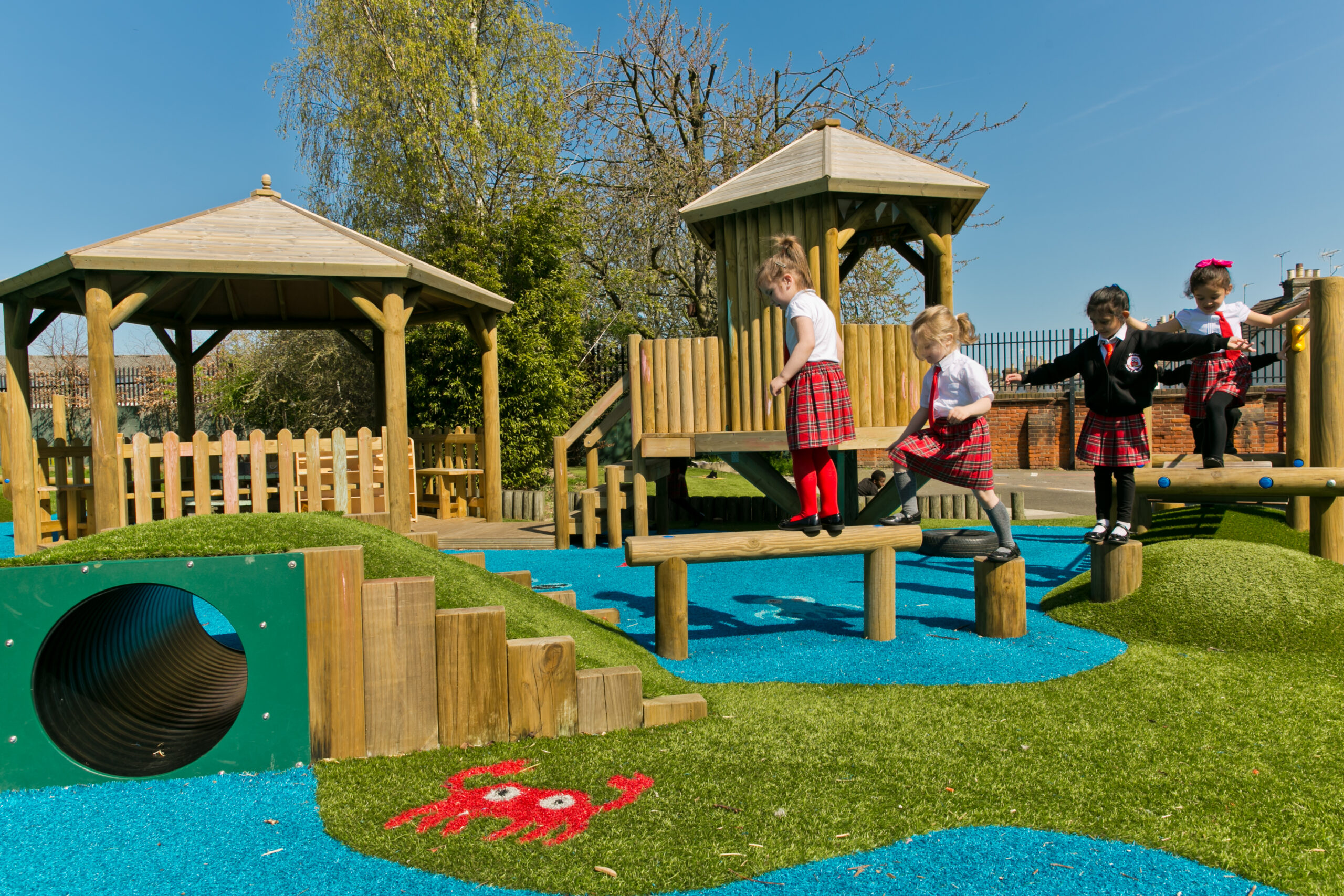Four young children in school uniforms play on a wooden playground structure with artificial grass and blue padding on a sunny day.