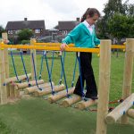 A girl in a turquoise jacket walks across a wooden and rope balance bridge on a playground in a grassy outdoor area.