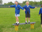 Two young girls in school uniforms stand balancing on a playground spring rocker in a grassy field, arms outstretched and smiling.