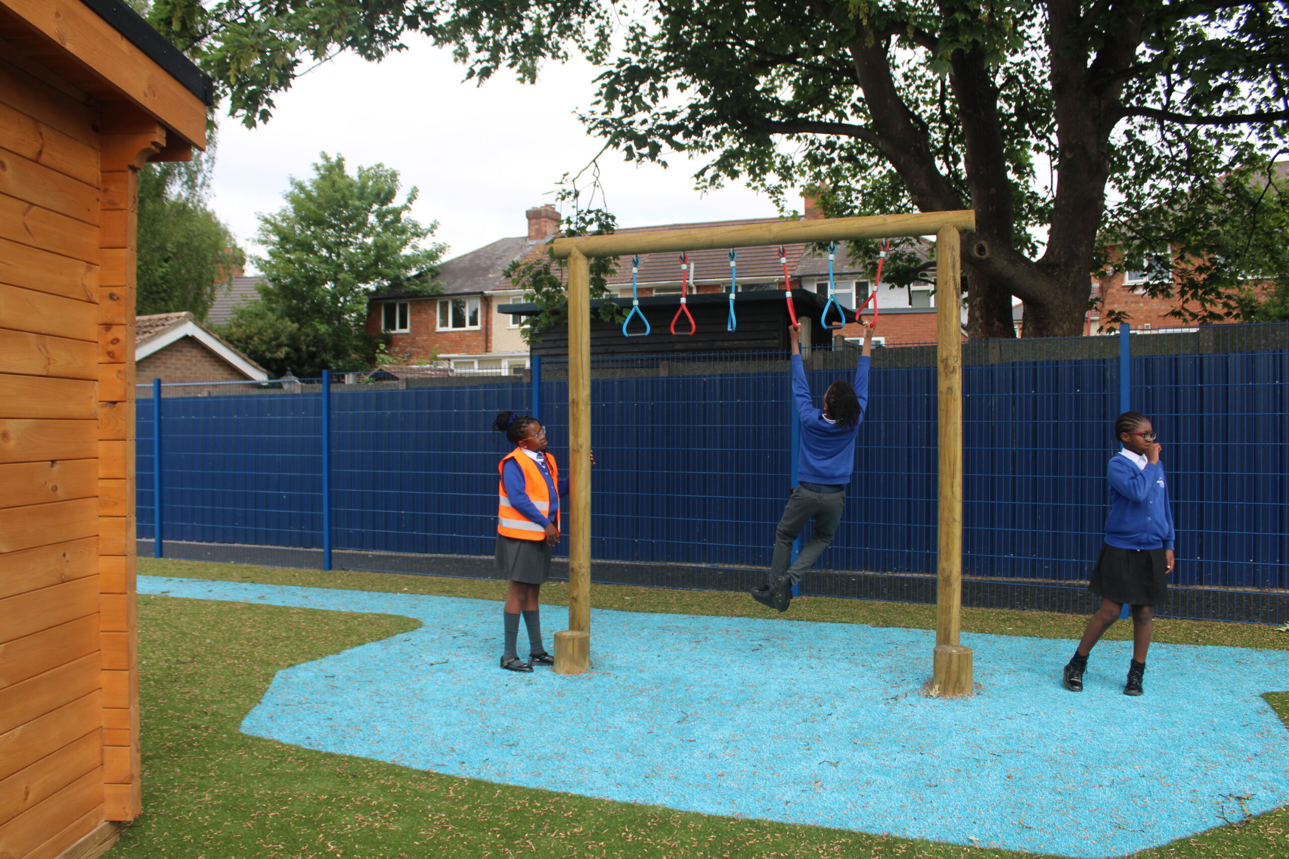 Three children in school uniforms are in a playground; one is on the Single Trapeze Swing, another stands nearby, and a third is using a mobile.