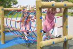 Two young girls in red chequered dresses play on a rope tunnel climbing structure at an outdoor playground on a sunny day.