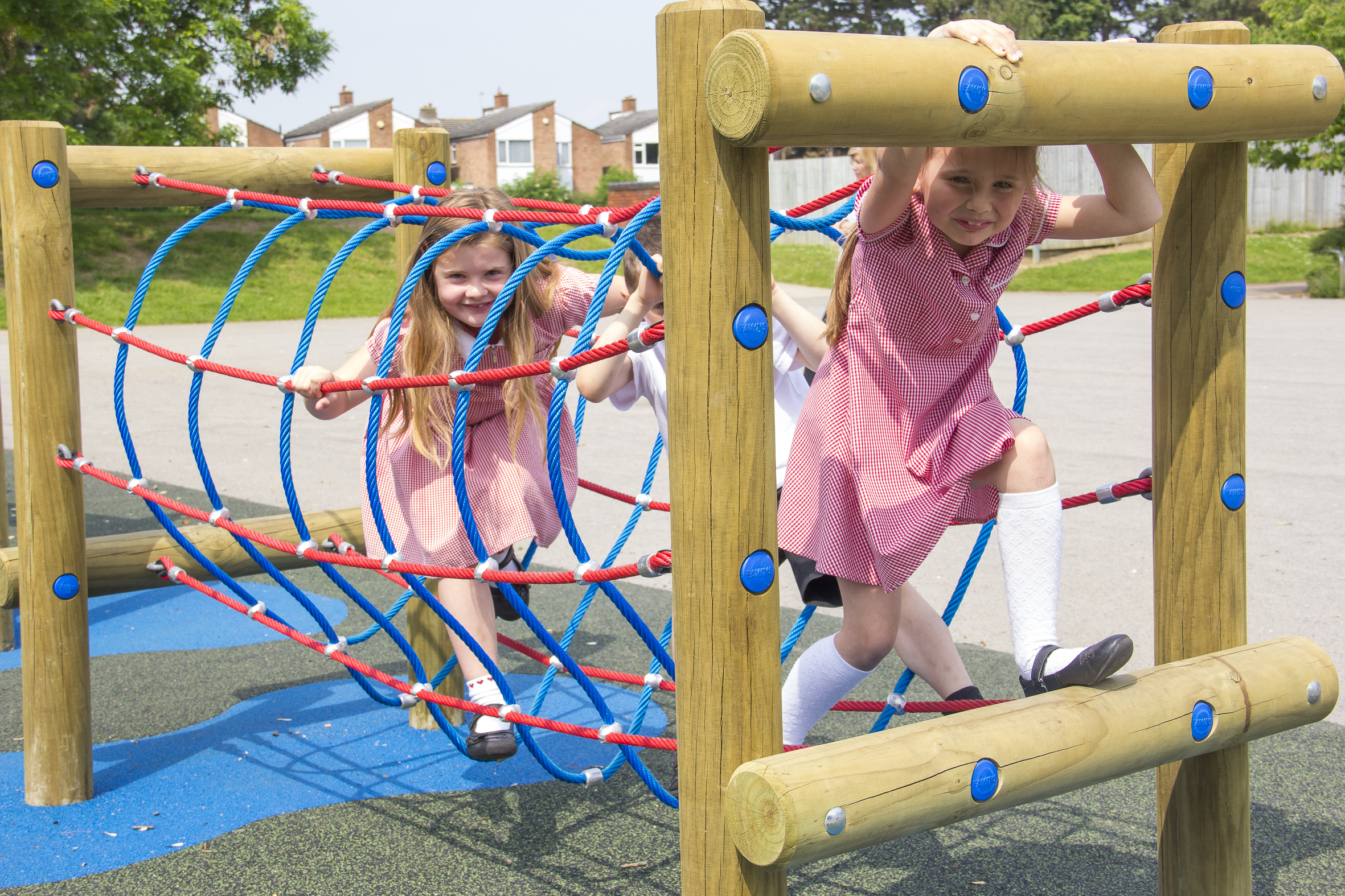 Two young girls in red chequered dresses play on a rope tunnel climbing structure at an outdoor playground on a sunny day.