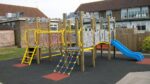 Outdoor playground set featuring the Excalibur Tower 600mm with Poly Slide, climbing nets, ladders, and platforms on a black safety surface in front of a school building.