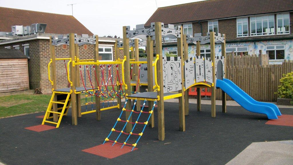 Outdoor playground set featuring the Excalibur Tower 600mm with Poly Slide, climbing nets, ladders, and platforms on a black safety surface in front of a school building.