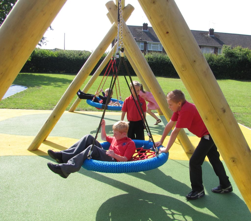 Children in red shirts play on large rope swings in a playground with wooden frames, artificial grass, and nearby houses in the background.