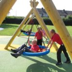 Children in red shirts play on large rope swings in a playground with wooden frames, artificial grass, and nearby houses in the background.