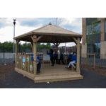 Children in school uniforms sit and stand inside a wooden outdoor gazebo on a school playground, with a fence and building in the background.