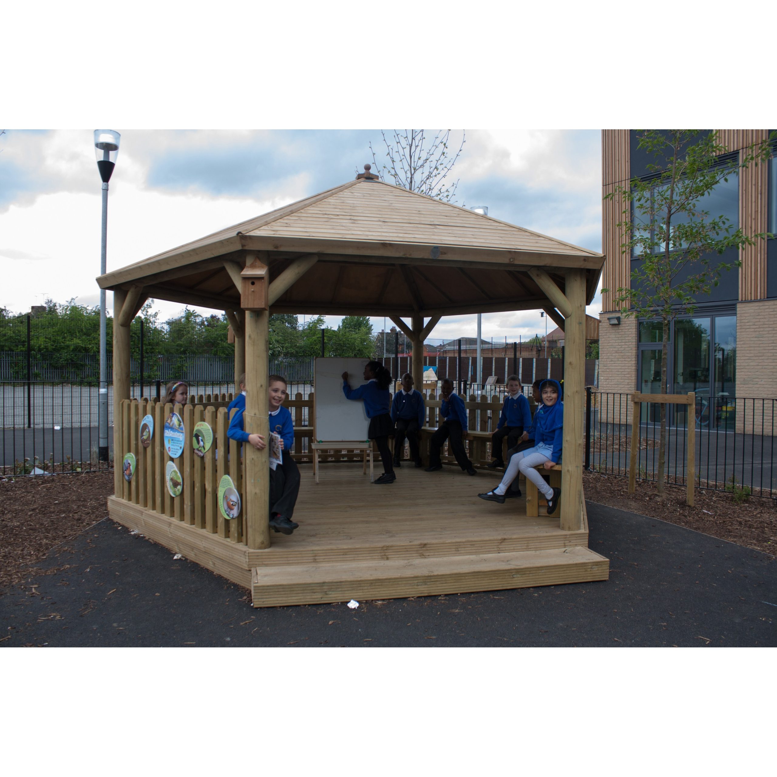Children in school uniforms sit and stand inside a wooden outdoor gazebo on a school playground, with a fence and building in the background.