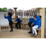 A group of schoolchildren in blue uniforms sit on benches outdoors whilst one girl writes on a whiteboard.