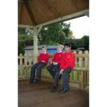 Three boys in red school uniforms sit on a wooden bench under a gazebo in an outdoor setting, with a wooden fence and greenery in the background.