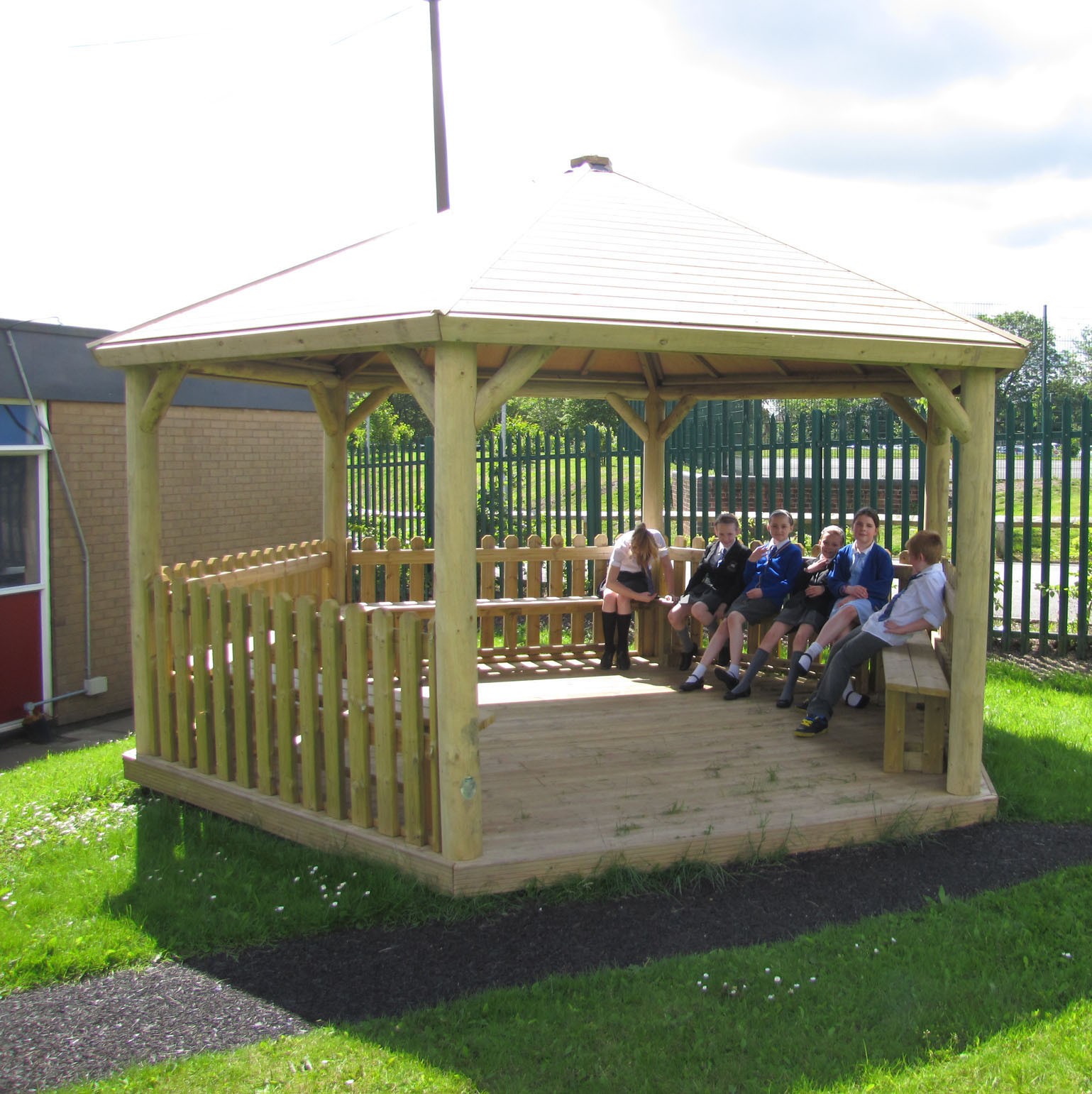 A group of children sitting on benches inside a wooden gazebo on a grassy area near a building and a metal fence.