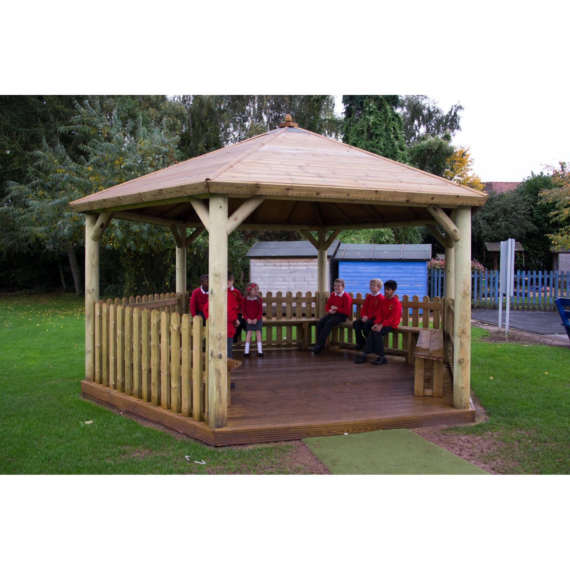 Four children in red uniforms sit inside a wooden gazebo with a picket fence, situated on a grassy area with trees and playground equipment in the background.