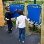 Two young boys stand at outdoor blue blackboards, one writing whilst the other watches, on a playground with a fence and grassy area in the background.