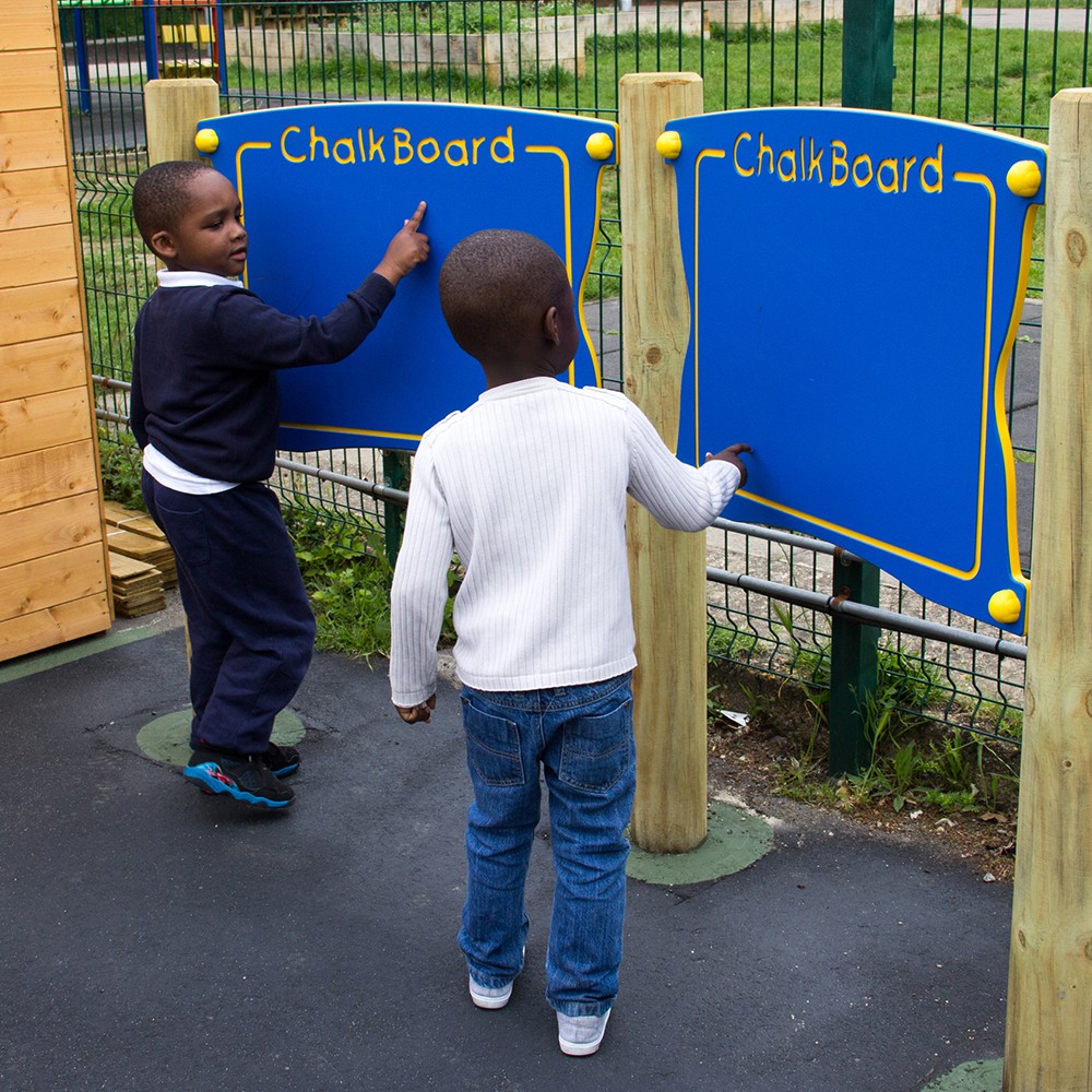 Two young boys stand at outdoor blue blackboards, one writing whilst the other watches, on a playground with a fence and grassy area in the background.