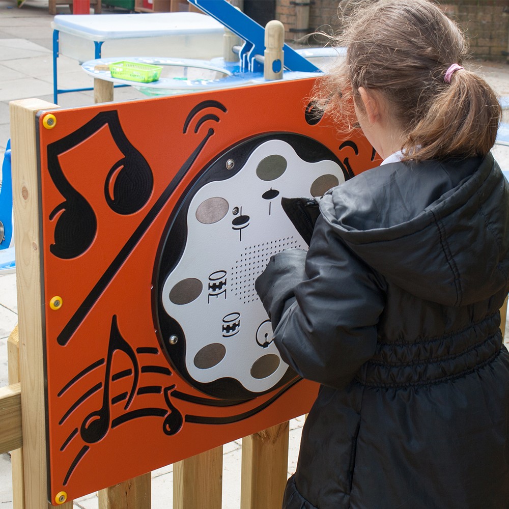 A child in a black jacket interacts with an outdoor musical play panel featuring black musical notes and symbol designs.