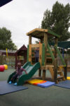 Two young children play on a Young Explorer – Crusoe 900mm wooden playground structure; one slides down a green slide whilst the other stands at the top platform. Outdoor setting with trees and play equipment visible.