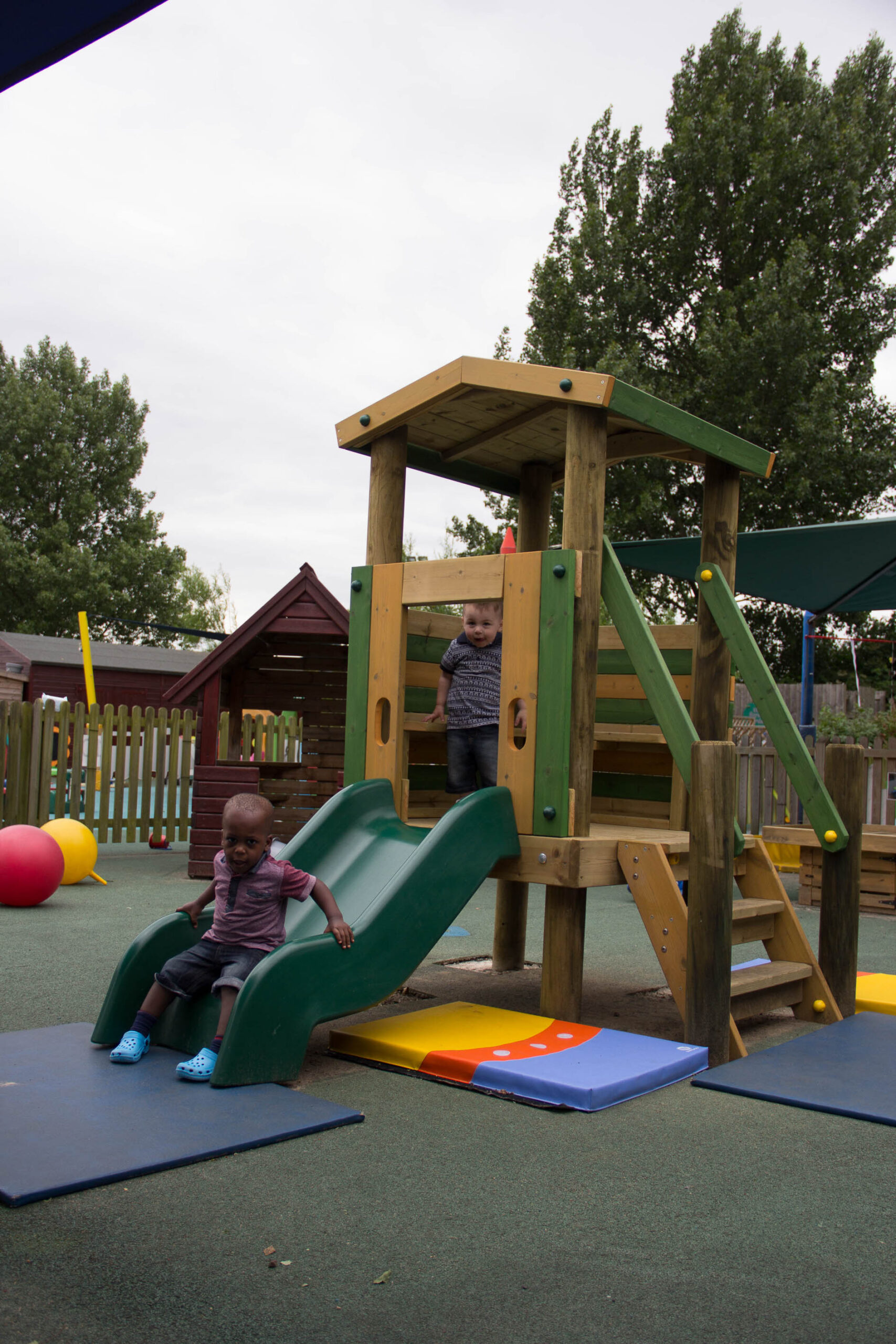 Two young children play on a Young Explorer – Crusoe 900mm wooden playground structure; one slides down a green slide whilst the other stands at the top platform. Outdoor setting with trees and play equipment visible.