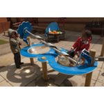 Two children play at an outdoor water table with a large metal screw mechanism, surrounded by playground equipment and toys.