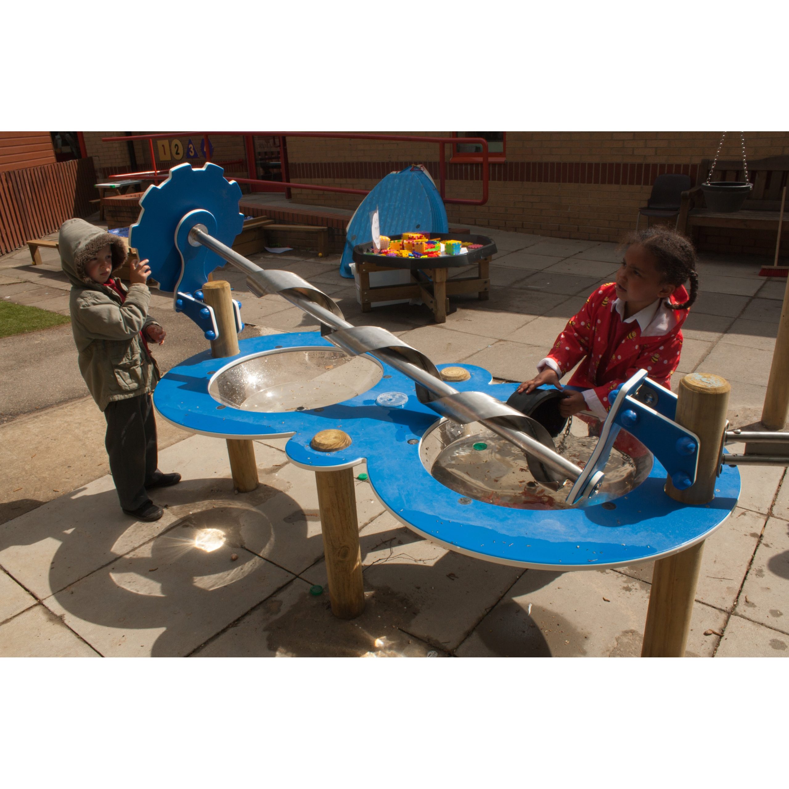 Two children play at an outdoor water table with a large metal screw mechanism, surrounded by playground equipment and toys.