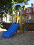 A children’s playground structure featuring the Ariane 900mm with Poly Deck Slide and a climbing net, set on wood chippings, with a brick building and fence in the background.