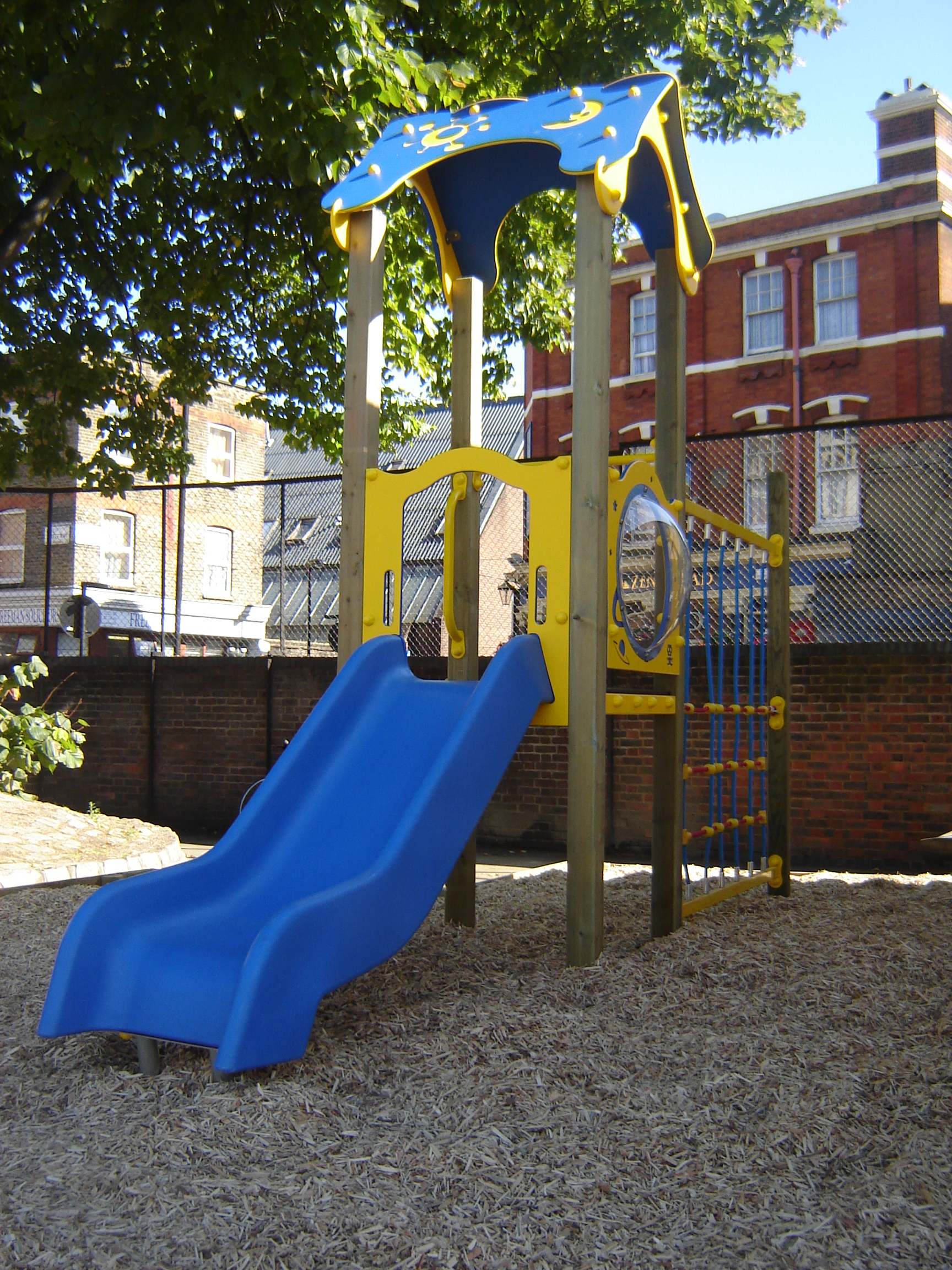 A children’s playground structure featuring the Ariane 900mm with Poly Deck Slide and a climbing net, set on wood chippings, with a brick building and fence in the background.